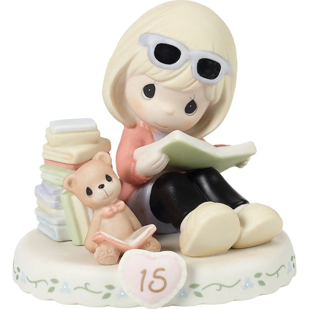 A Precious Moments figurine of a girl with short blonde hair and white sunglasses on her head, sitting and reading a book next to a teddy bear and a stack of books on a decorative base with a heart that says '15'.