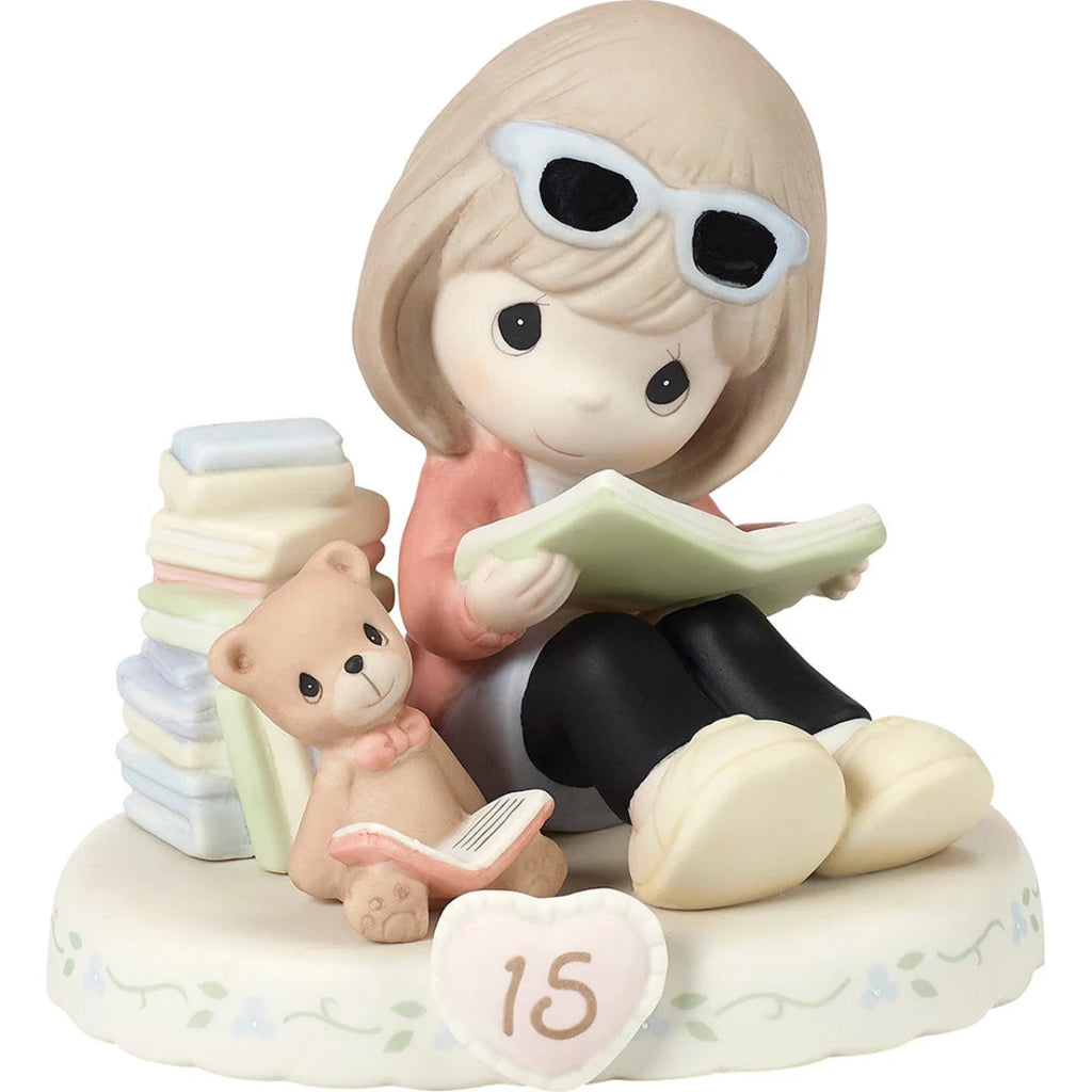 A ceramic figurine of a young girl with short brunette hair and white sunglasses on her head sitting on a base, reading a book next to a teddy bear and a stack of books on a decorative base with a heart that says '15'.