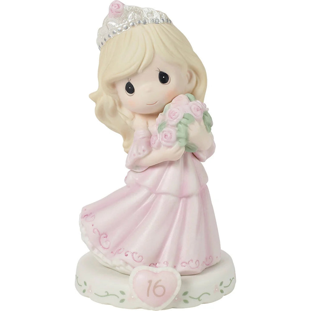 A Precious Moments figurine of a young girl with blonde hair wearing a tiara and a pink dress, holding a bouquet of pink roses. The base features a heart with the number 16.
