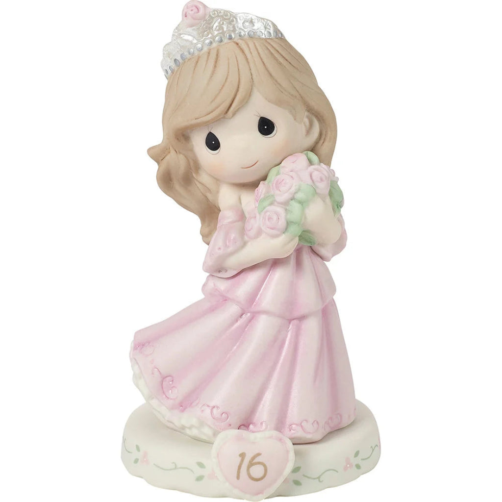 A Precious Moments figurine of a girl  with brunette hair celebrating her 16th birthday, wearing a pink gown and tiara while holding a bouquet of pink roses, set on a base with a heart-shaped plaque displaying the number 16.
